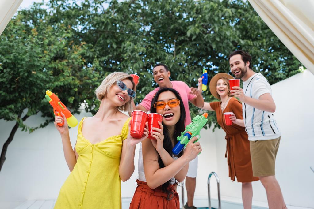 různé ženy s vodní pistole smích a opékání s plastovými poháry v blízkosti šťastných přátel - Fotografie, Obrázek