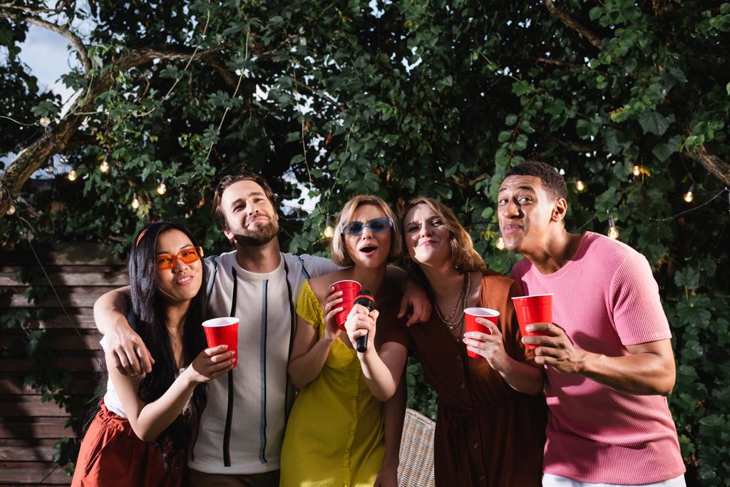 allegri amici multietnici in possesso di bicchieri di plastica mentre cantano karaoke in giardino di notte - Foto, immagini