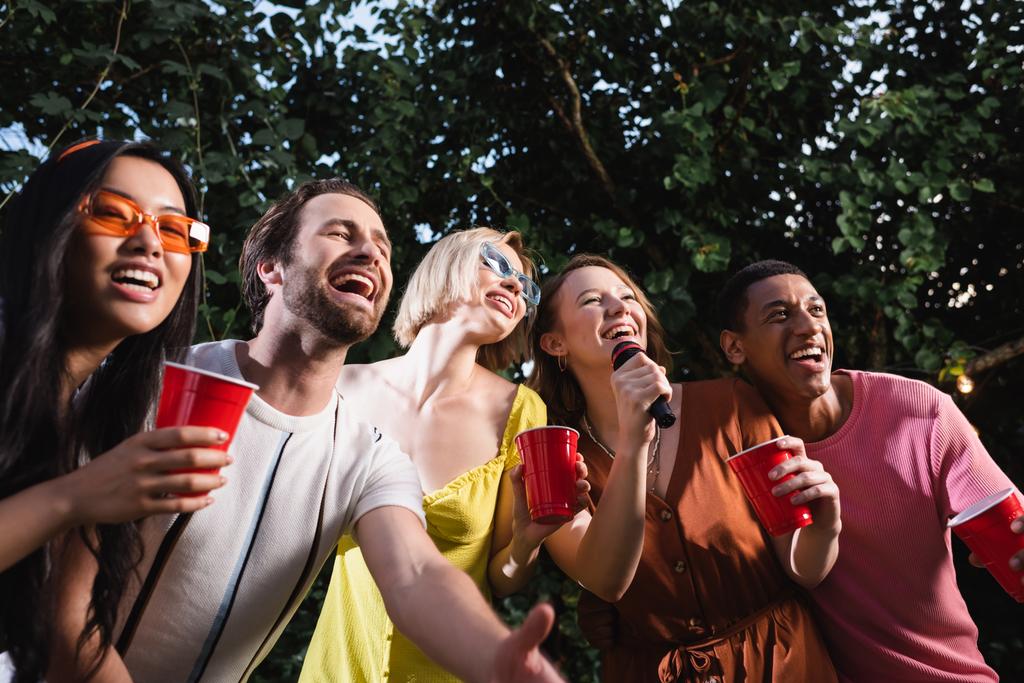 Pessoas multiétnicas alegres cantando karaoke e segurando bebida em copos de plástico ao ar livre  - Foto, Imagem