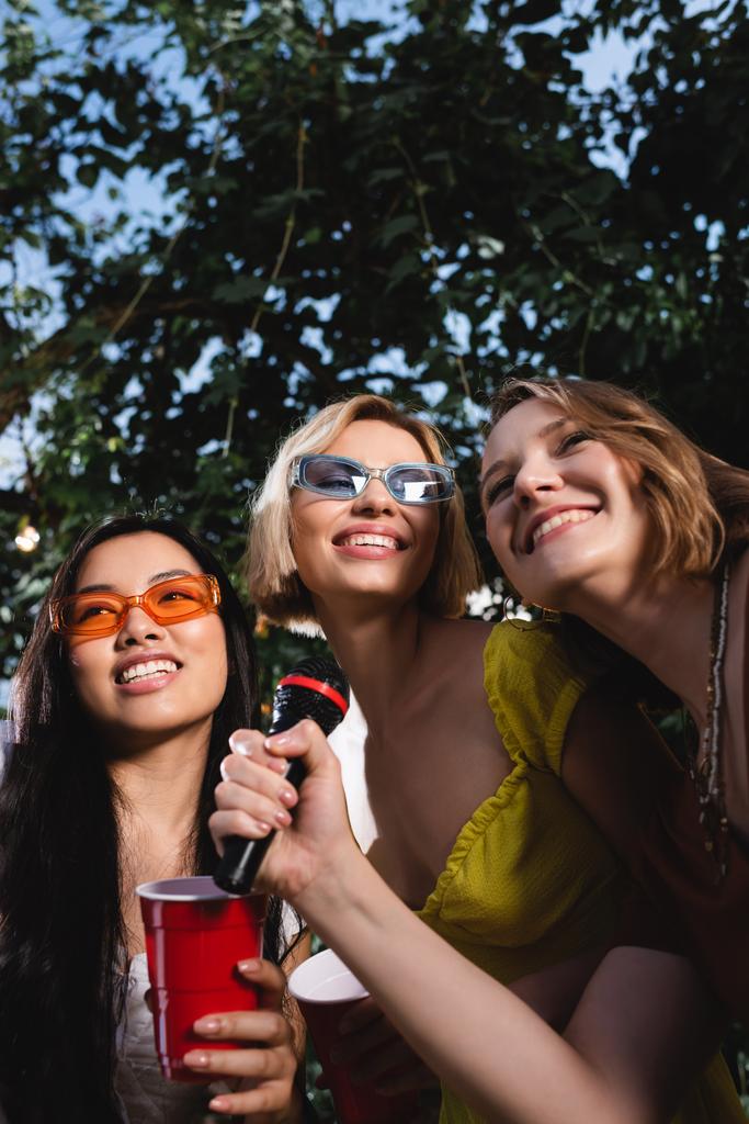 Vista de ángulo bajo de mujeres multiétnicas sonrientes con taza de plástico que sostiene el micrófono al aire libre durante la fiesta  - Foto, imagen