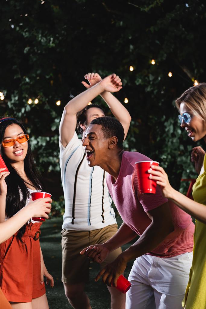 Positive african american man dancing with interracial friends holding plastic cups during summer party  - Foto, afbeelding