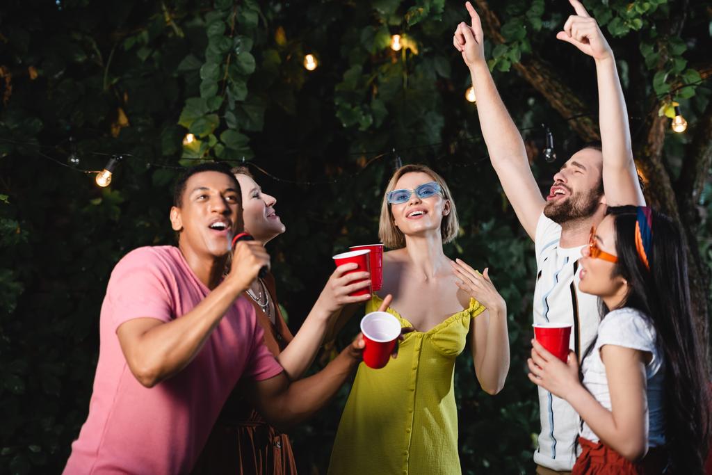 Uomo afroamericano che canta karaoke vicino agli amici con bicchieri di plastica all'aperto  - Foto, immagini