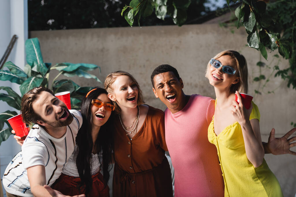 Amigos multiétnicos alegres com copos de plástico se divertindo ao ar livre  - Foto, Imagem