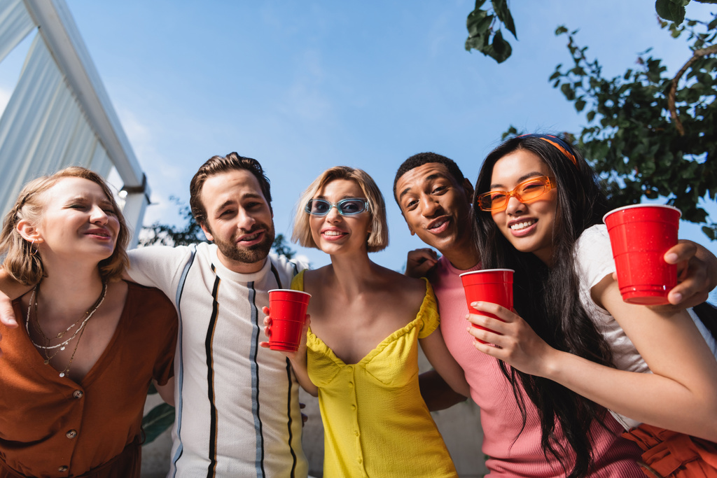 Vue à angle bas d'amis multiculturels avec boisson regardant la caméra pendant la fête  - Photo, image