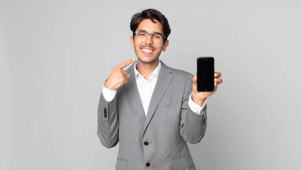 joven hombre hispano sonriendo con confianza señalando a su propia sonrisa amplia y sosteniendo un teléfono inteligente - Foto, Imagen