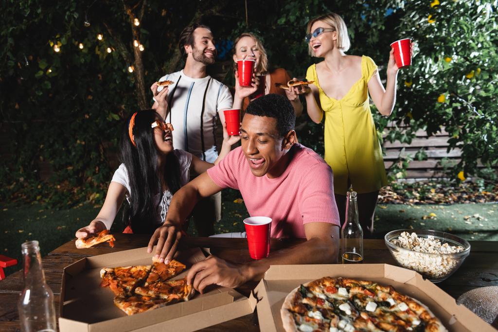 Afro-Amerikalı bir adam gülümseyerek arkadaşlarının yanına pizza ve açık havada bira götürüyor.  - Fotoğraf, Görsel