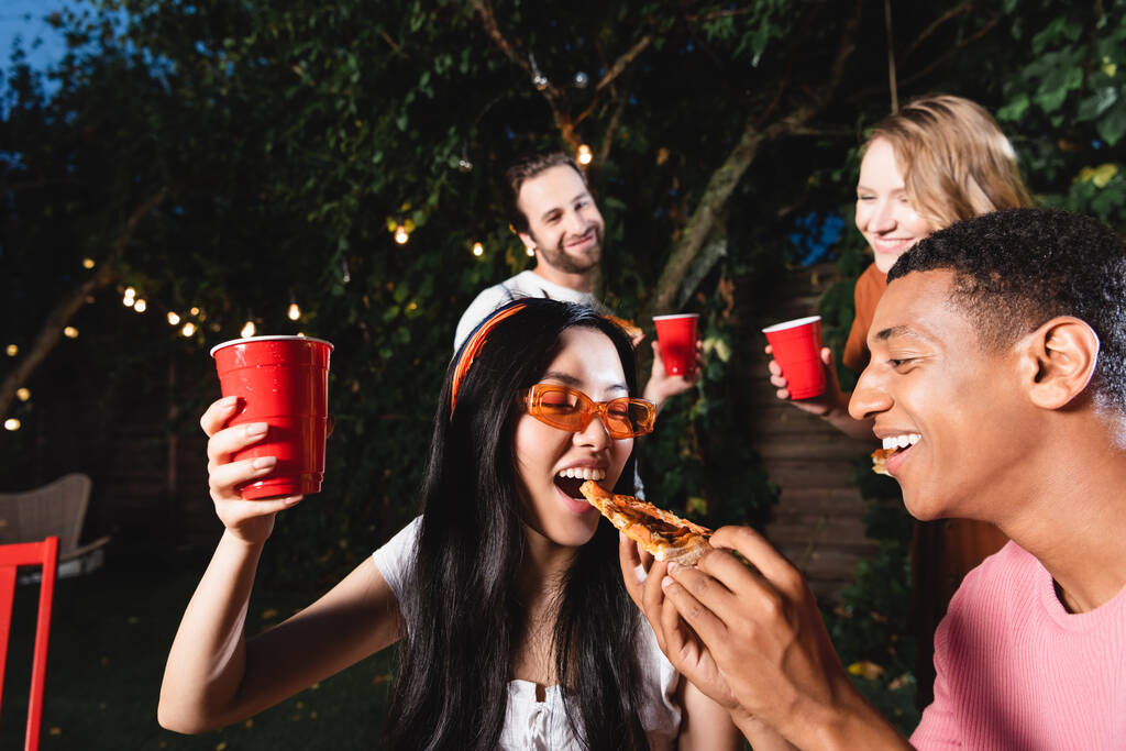 African american man feeding pizza to asian friend with plastic cup outdoors  - Zdjęcie, obraz