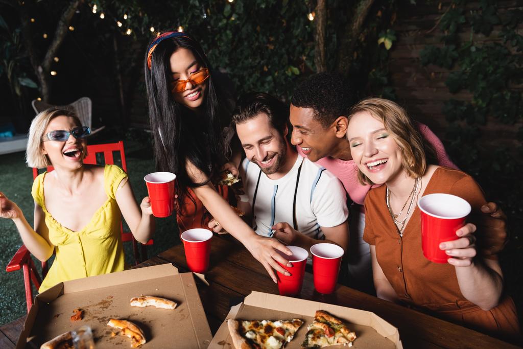 Laughing woman in sunglasses holding plastic cup near multiethnic friends and food outdoors  - Photo, Image