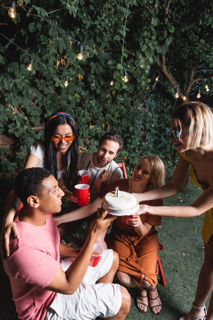 Веселые межрасовые друзья с пластиковыми чашками, празднующие день рождения африканского американца на открытом воздухе  - Фото, изображение