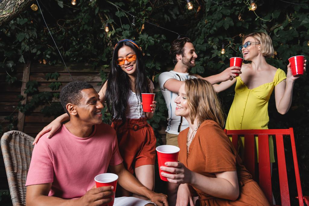 Jóvenes amigos interracial con copas de plástico hablando durante la fiesta de verano por la noche  - Foto, Imagen