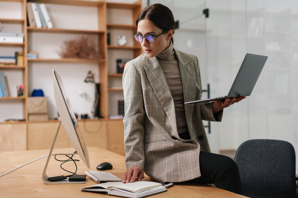 Focalisé femme charmante dans les lunettes de travail avec ordinateur et ordinateur portable au bureau - Photo, image