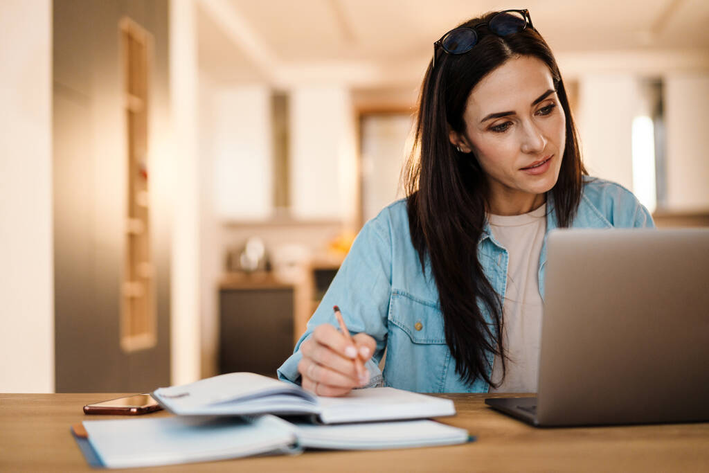 Konzentrierte, charmante Frau schreibt Notizen, während sie zu Hause mit Laptop arbeitet - Foto, Bild