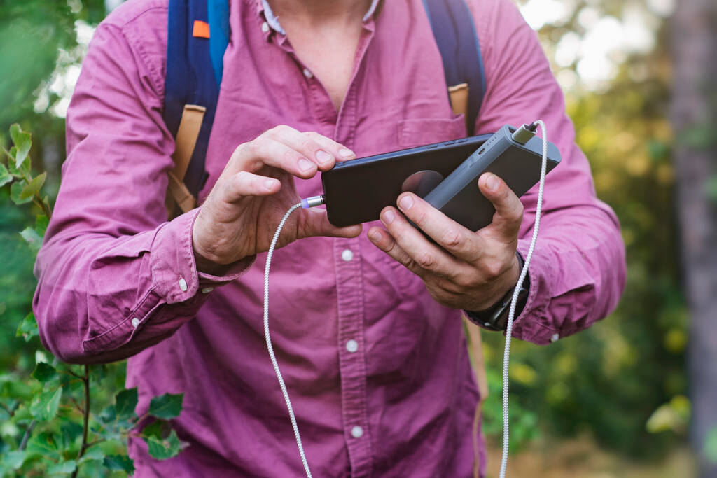 Turysta trzyma smartfona z przenośną ładowarką w rękach. Człowiek z Power Bankiem ładuje telefon na tle natury i lasu - Zdjęcie, obraz