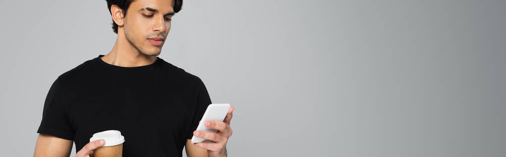 Молодой человек держит кофе идти и с помощью смартфона изолированы на серый, баннер - Фото, изображение