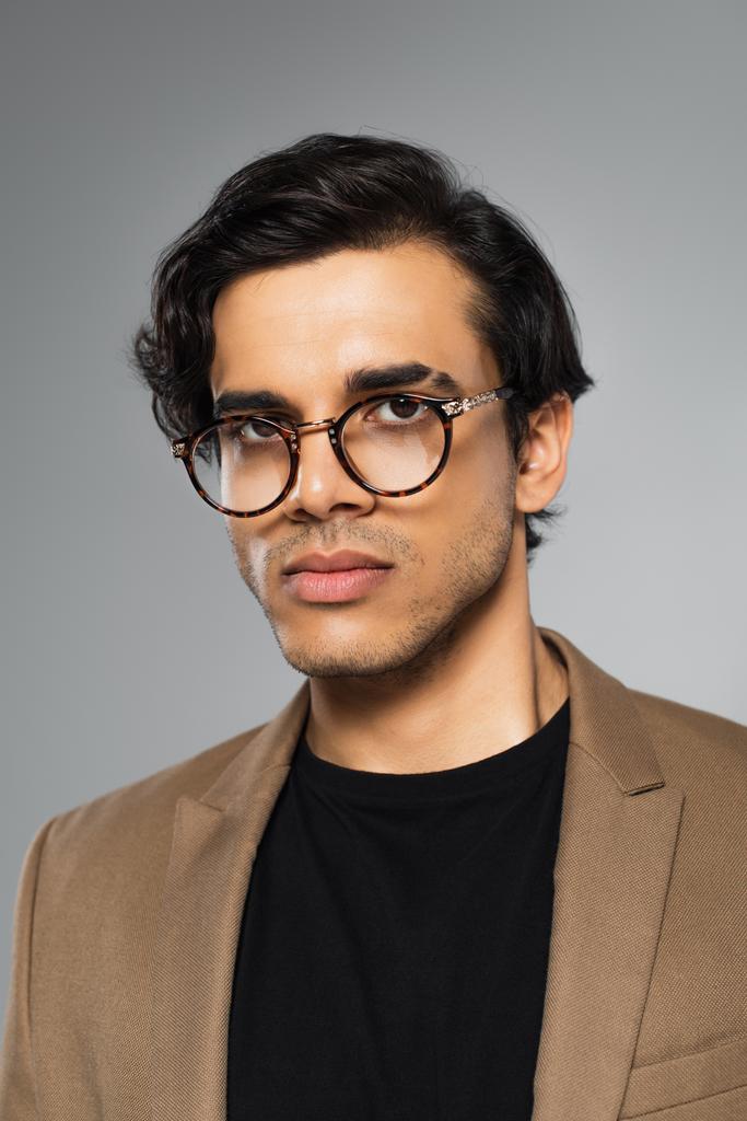 молодой человек в модных очках, смотрящий в камеру, изолированную на сером  - Фото, изображение