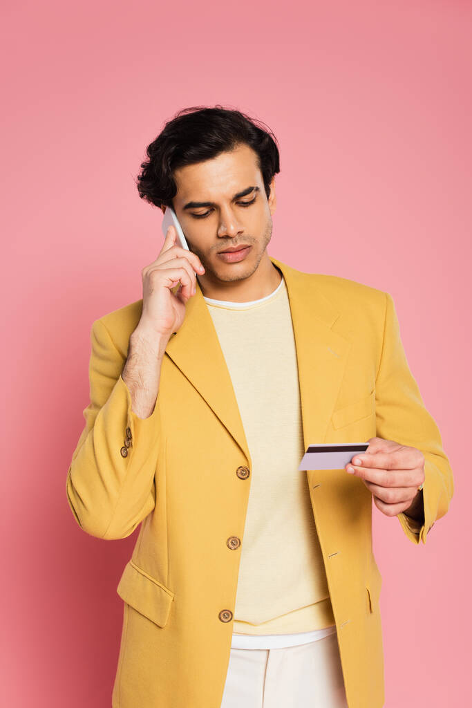 homem em blazer amarelo segurando cartão de crédito enquanto fala no smartphone isolado em rosa - Foto, Imagem