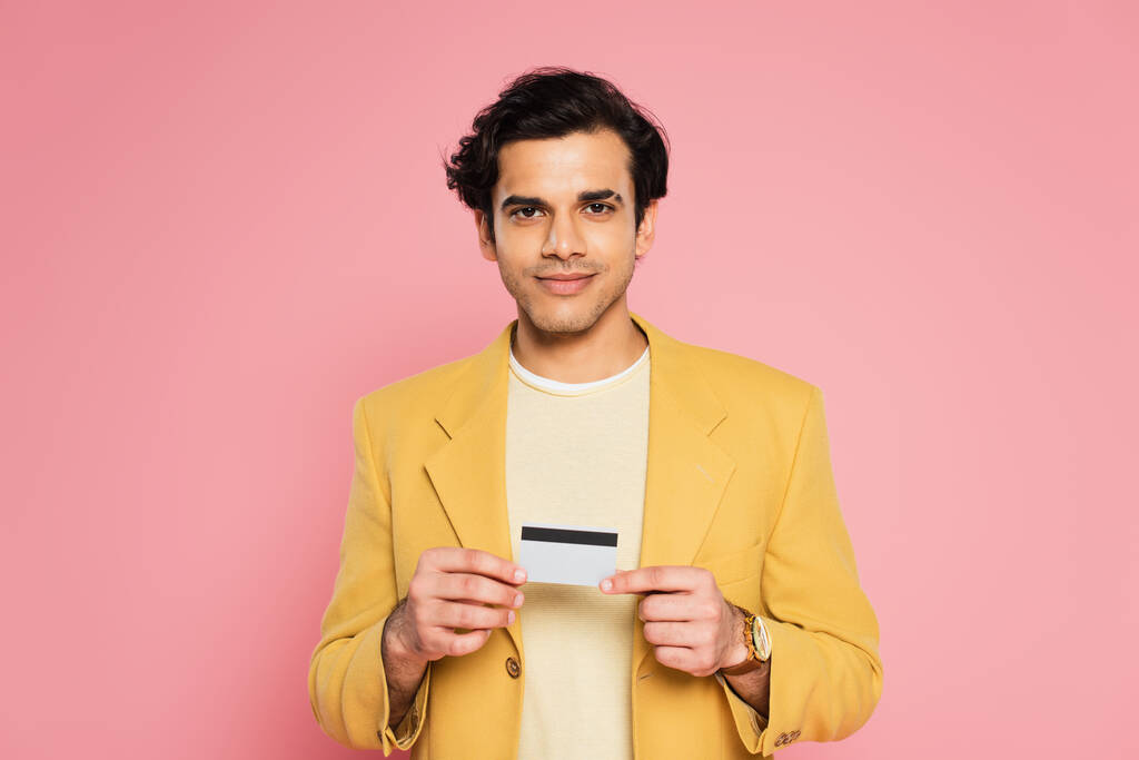 hombre sonriente en chaqueta amarilla con tarjeta de crédito aislada en rosa - Foto, imagen