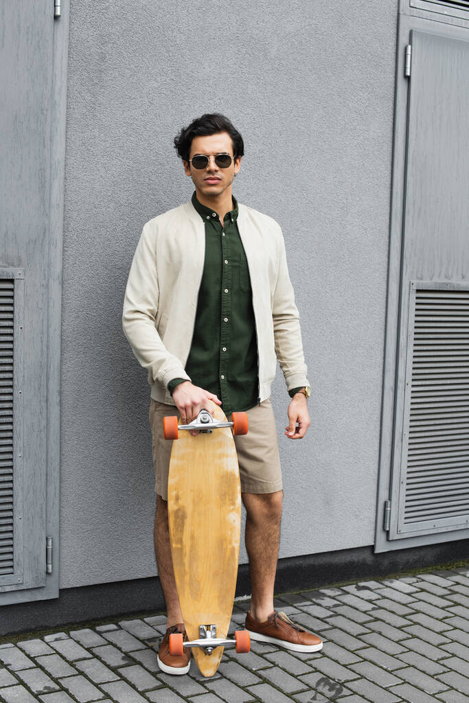 Stilvoller Mann mit Sonnenbrille hält Longboard in Baunähe  - Foto, Bild