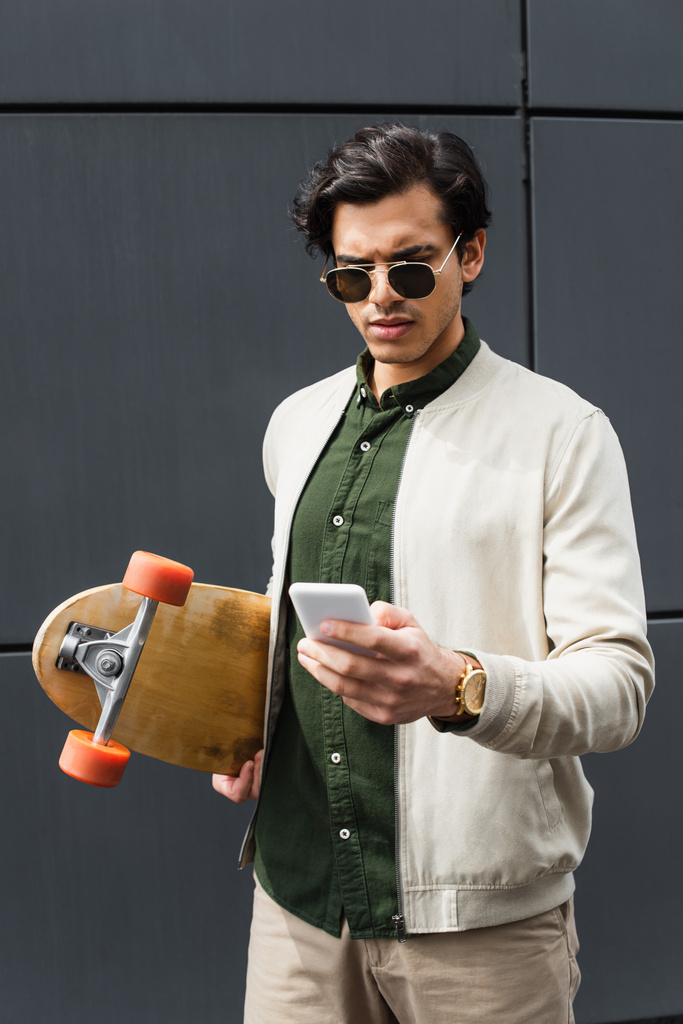 stylowy młody człowiek w okularach przeciwsłonecznych i bombowej kurtce trzymając longboard podczas pisania na smartfonie w pobliżu budynku  - Zdjęcie, obraz