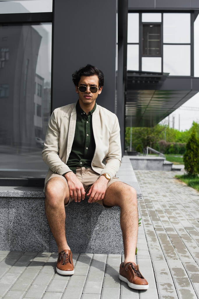longitud completa de hombre con estilo en gafas de sol sentado fuera  - Foto, imagen