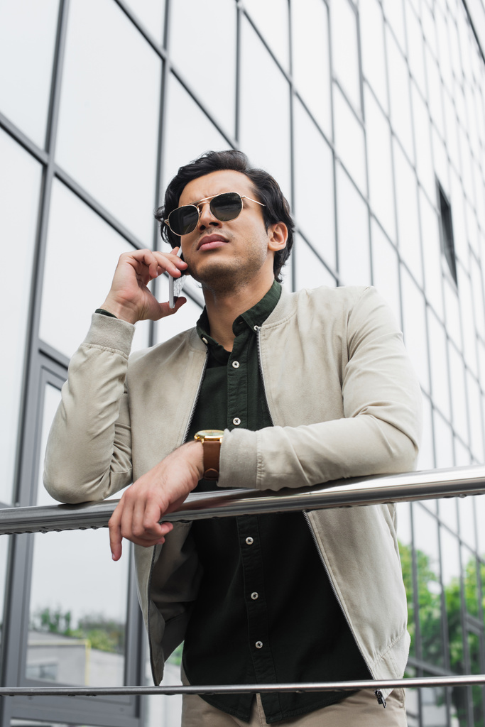 vista angolo basso di elegante giovane uomo in occhiali da sole che parla sul telefono cellulare  - Foto, immagini