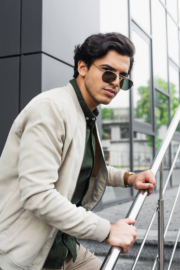 jeune homme avec des lunettes de soleil élégantes et une veste de bombardier posant près du bâtiment  - Photo, image