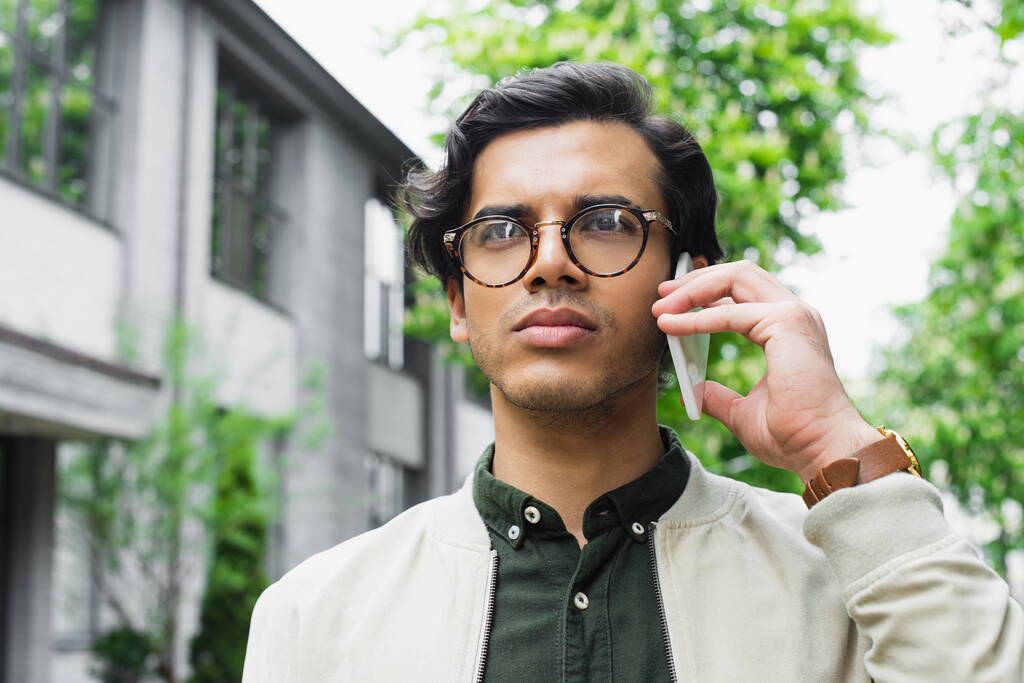 Junger Mann mit Brille und Bomberjacke schaut weg, während er draußen auf dem Smartphone spricht  - Foto, Bild