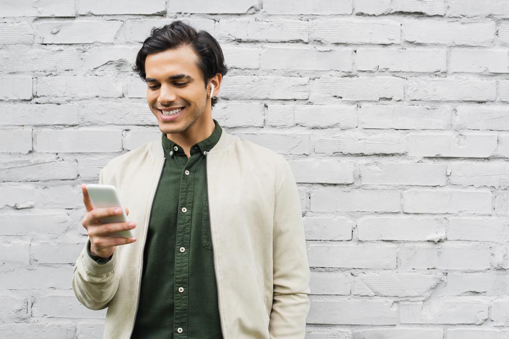fröhlicher junger Mann mit drahtlosen Kopfhörern lächelt, während er sein Smartphone in der Nähe der Ziegelmauer benutzt - Foto, Bild