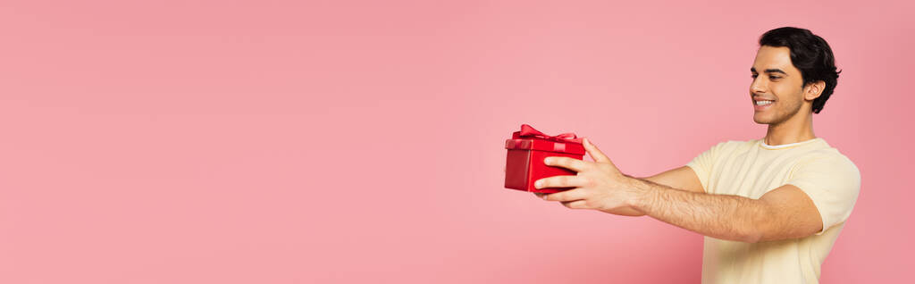 nuori iloinen mies tilalla punainen lahjapakkaus eristetty vaaleanpunainen, banneri - Valokuva, kuva