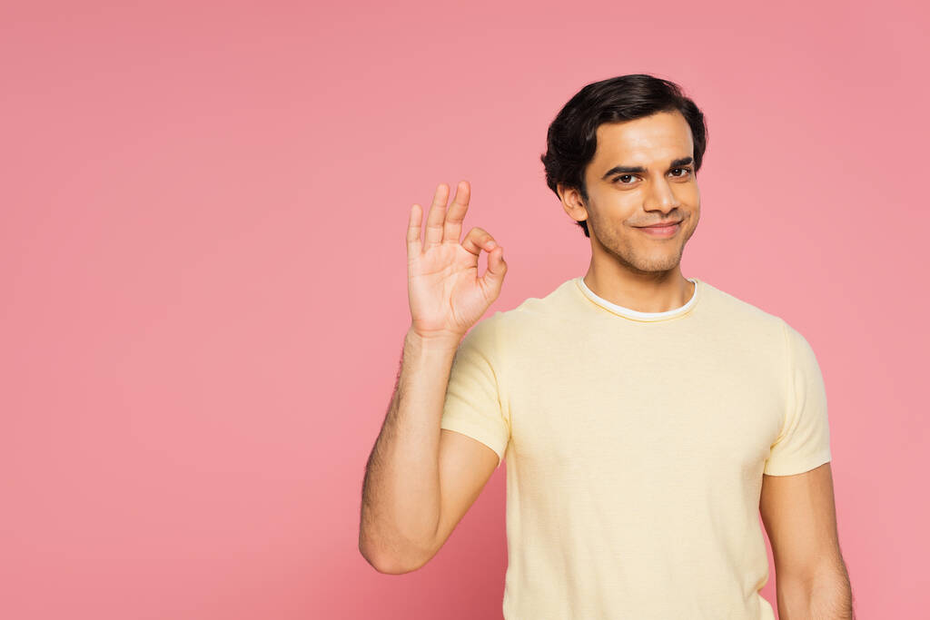 veselý mladý muž usmívá a ukazuje ok znamení izolované na růžové - Fotografie, Obrázek