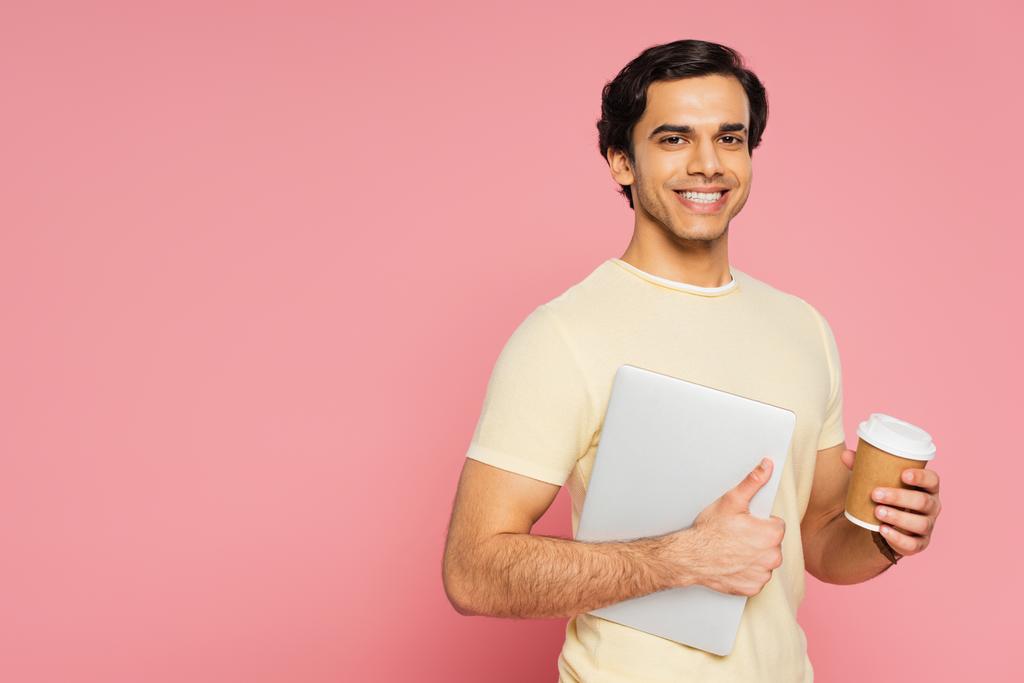 χαρούμενος ελεύθερος επαγγελματίας κρατώντας φορητό υπολογιστή και χάρτινο κύπελλο απομονώνονται σε ροζ - Φωτογραφία, εικόνα