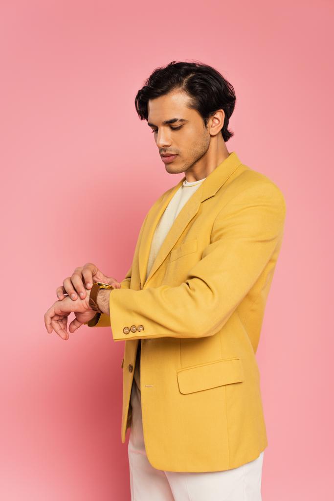κομψό νεαρό άνδρα σε μοντέρνο σακάκι κοιτάζοντας ρολόι χειρός σε ροζ  - Φωτογραφία, εικόνα