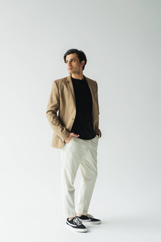 pleine longueur de jeune homme en élégant blazer beige debout avec les mains dans les poches sur le gris - Photo, image