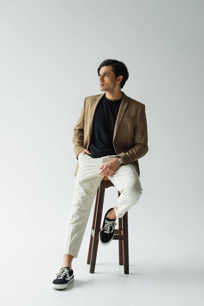 jonge man in stijlvolle beige blazer zittend op houten stoel op grijs - Foto, afbeelding