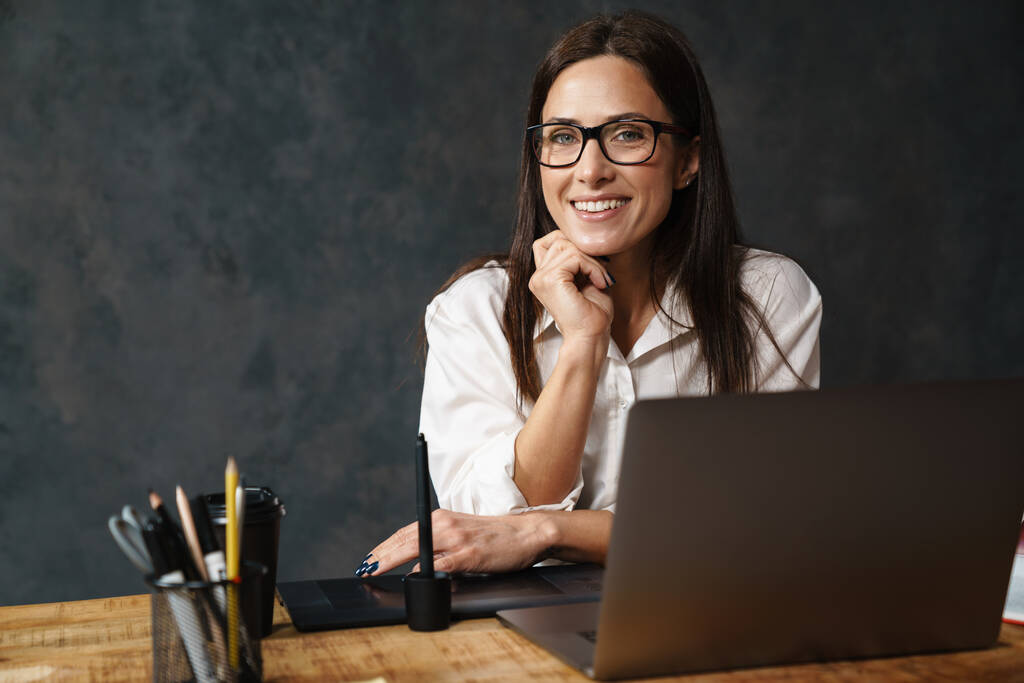 Uśmiechnięta brunetka w średnim wieku bizneswoman w koszuli siedzi przy biurku z laptopem na ciemnym tle - Zdjęcie, obraz