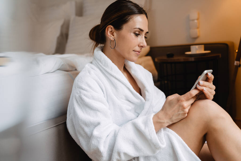 Усміхнена середнього віку брюнетка біла жінка в халаті сидить на підлозі в ліжку в приміщенні смс на мобільний телефон
 - Фото, зображення