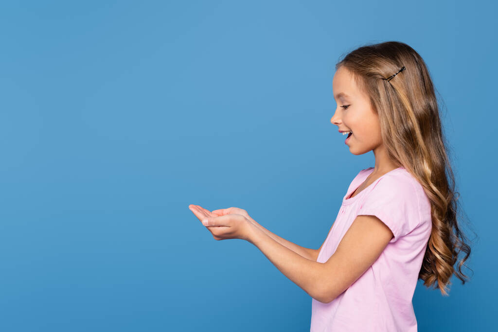 Vista lateral da menina sorridente com as mãos de chávena isoladas no azul - Foto, Imagem