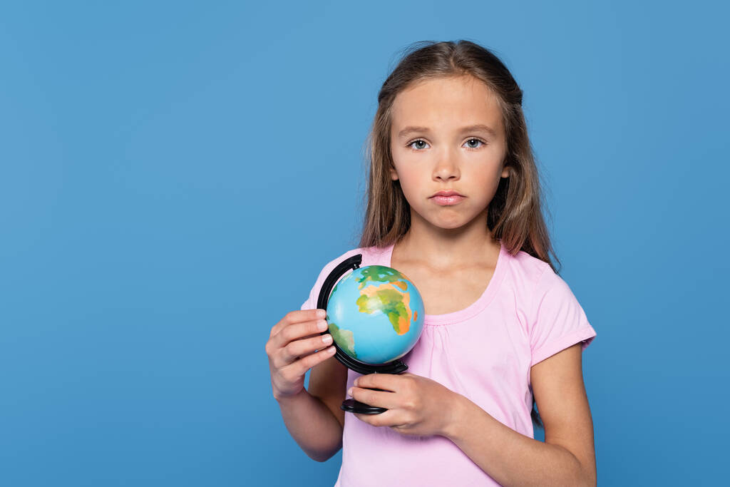 Zdenerwowany dzieciak z globusem patrzący na odizolowaną na niebiesko kamerę - Zdjęcie, obraz
