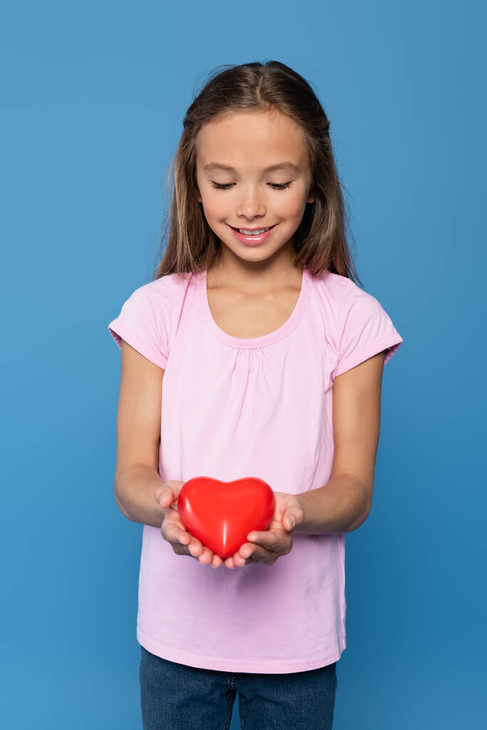 Smiling girl in pink t-shirt holding red heart isolated on blue - Photo, Image