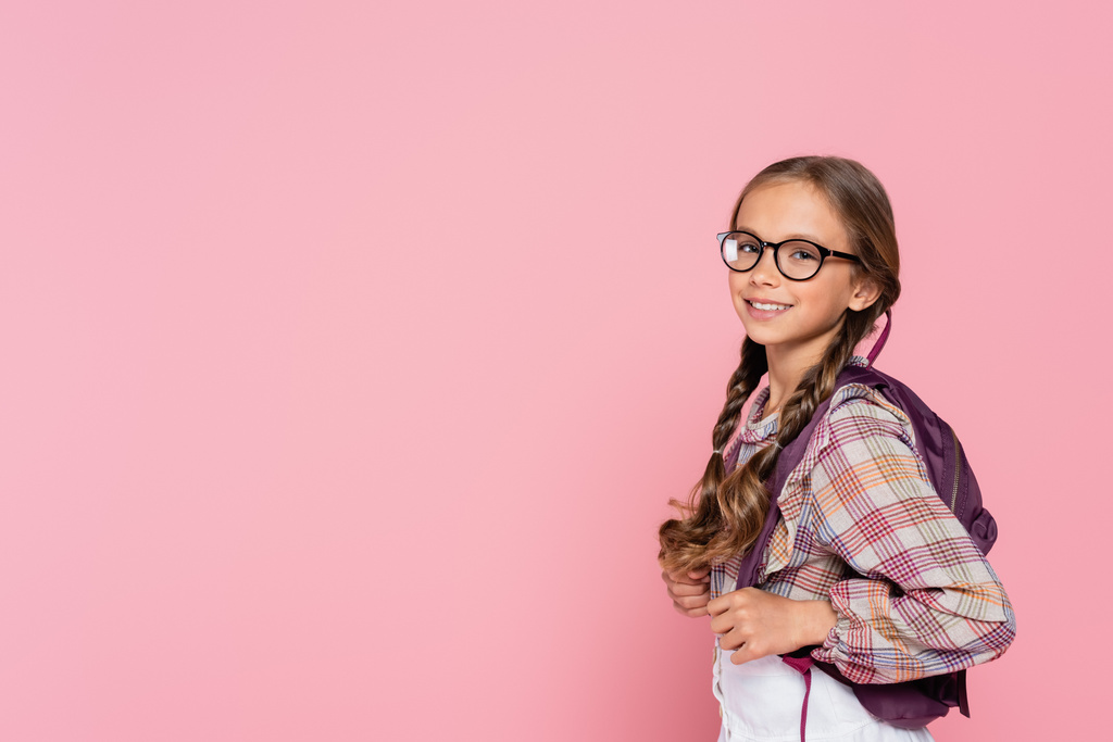 Preteen schoolgirl in eyeglasses and backpack smiling at camera isolated on pink  - Foto, imagen