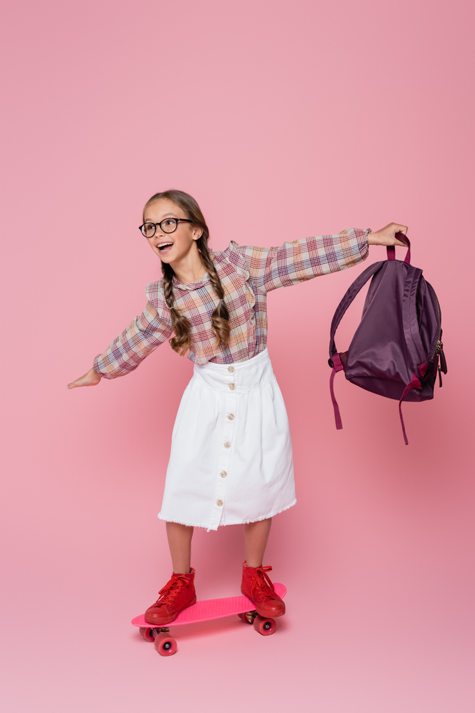 Schoolgirl with backpack riding penny board on pink background  - Foto, Imagen