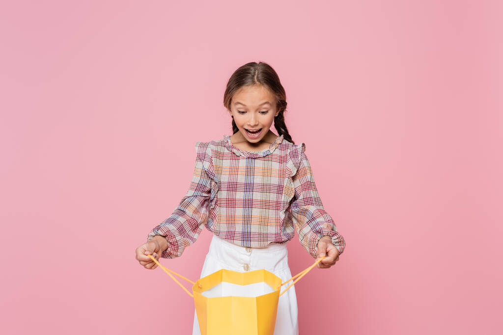 surprised child in plaid blouse looking into yellow shopping bag isolated on pink - Photo, Image