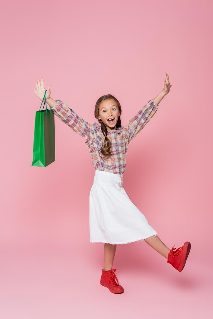 ενθουσιασμένοι κορίτσι με πράσινη τσάντα ψώνια σηκώνοντας τα χέρια σε ροζ φόντο - Φωτογραφία, εικόνα