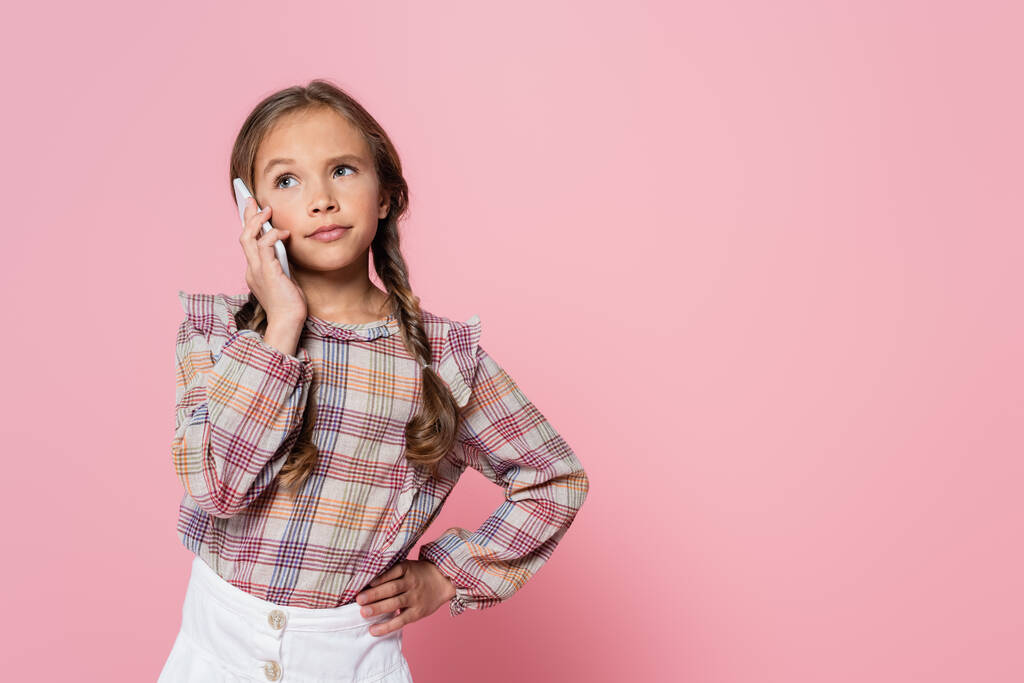 serious preteen girl talking on smartphone while standing with hand on hip isolated on pink - Foto, imagen