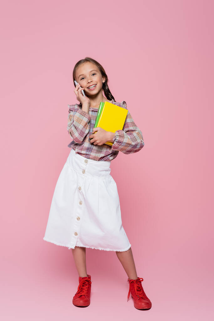 volledige weergave van trendy, vrolijk meisje met boeken praten op mobiele telefoon op roze achtergrond - Foto, afbeelding