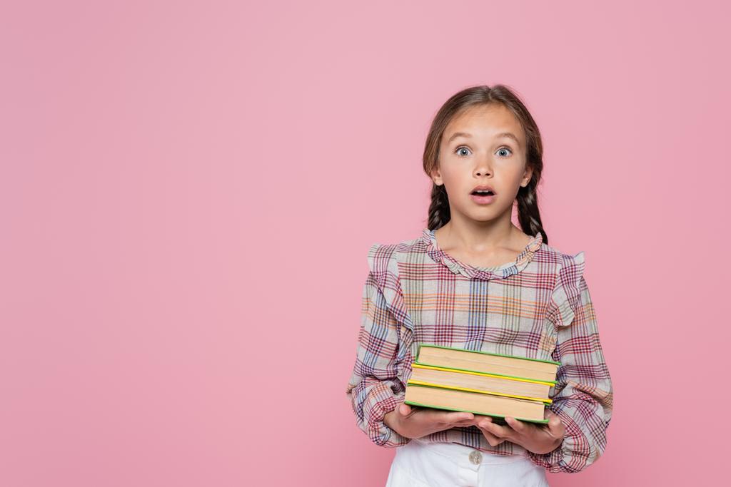 verrast kind op zoek naar camera terwijl het houden van boeken geïsoleerd op roze - Foto, afbeelding