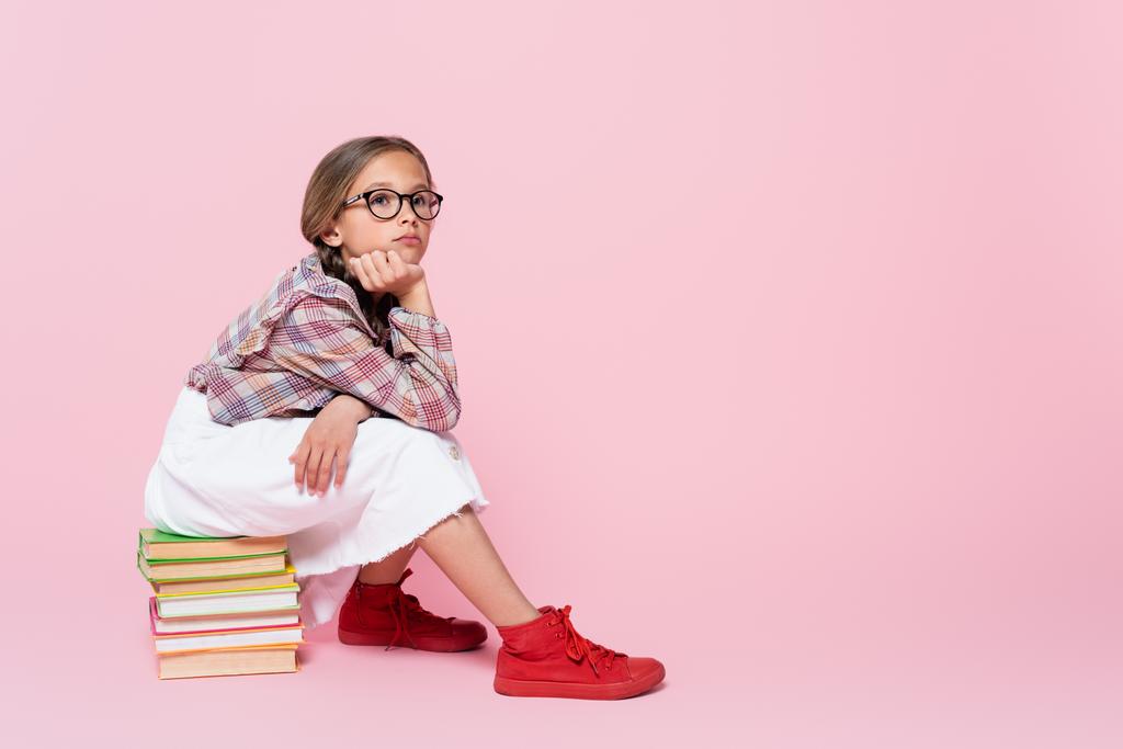 pensive girl in plaid blouse and white skirt sitting on books and looking away on pink background - Photo, Image