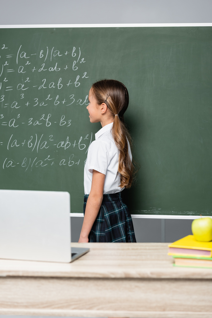 μαθήτρια στέκεται κοντά στο chalkboard με εξισώσεις και γραφείο με laptop  - Φωτογραφία, εικόνα