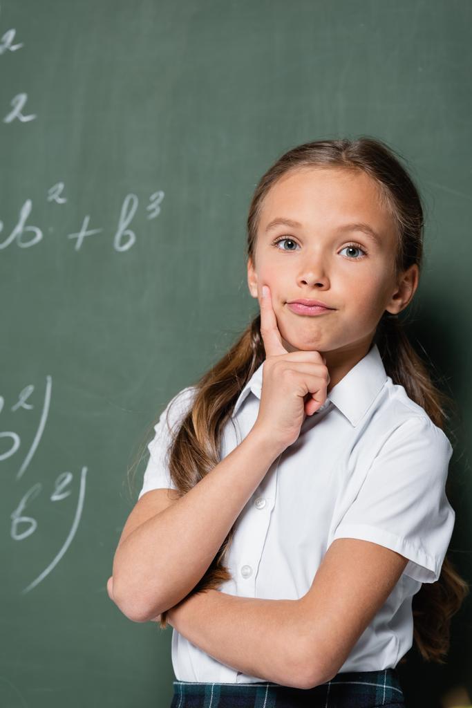 thoughtful schoolkid looking at camera near chalkboard in school - Photo, Image
