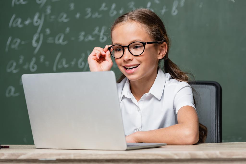 positive schoolkid adjusting eyeglasses near laptop and chalkboard on blurred background - Foto, Imagen